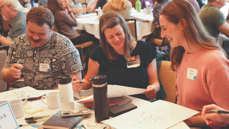 Group of people seated at a conference table, looking at papers and each other and smiling. The background shows many other people also at conference tables.