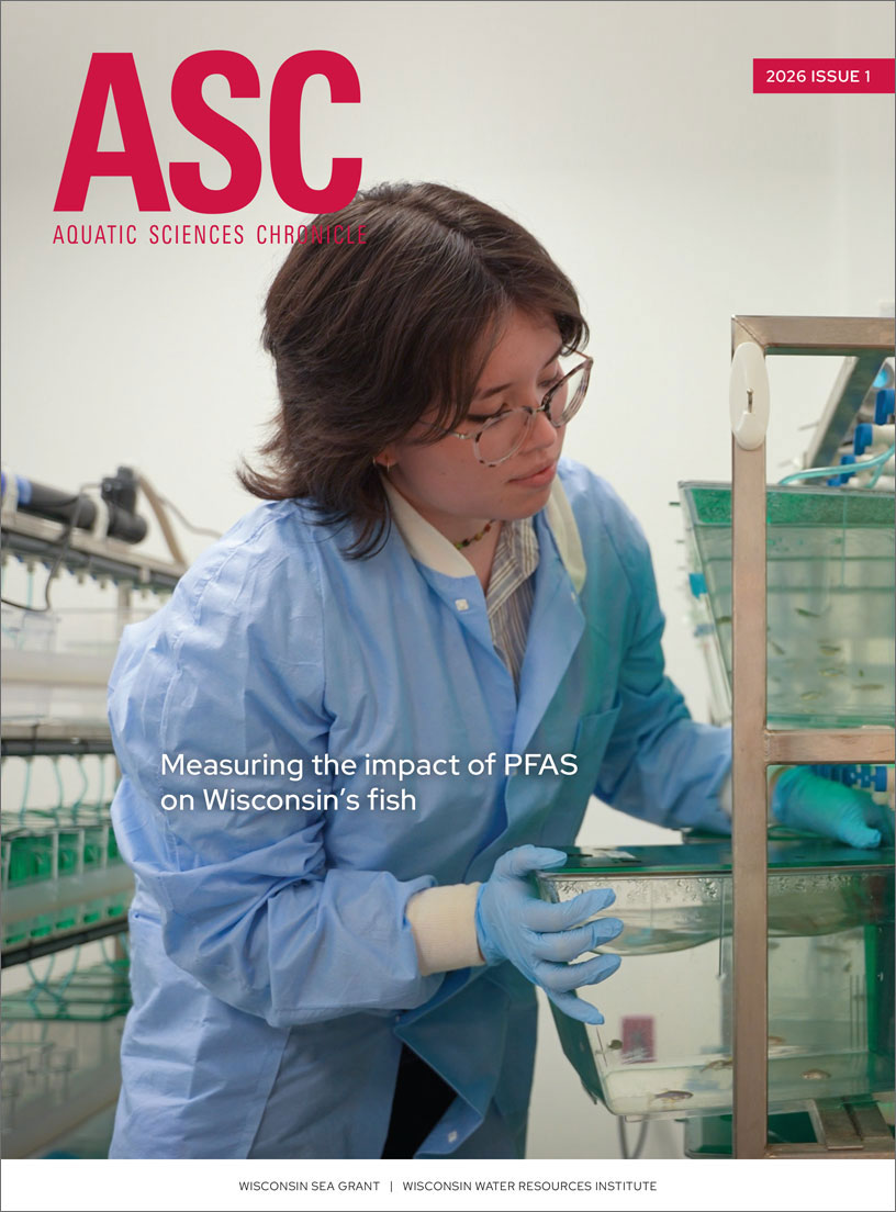 Aquatic Sciences Chronicle 2026 Issue 1 cover featuring a woman moving a small tank of fish into a rack of similar tanks with a row of still more fish tanks behind her.