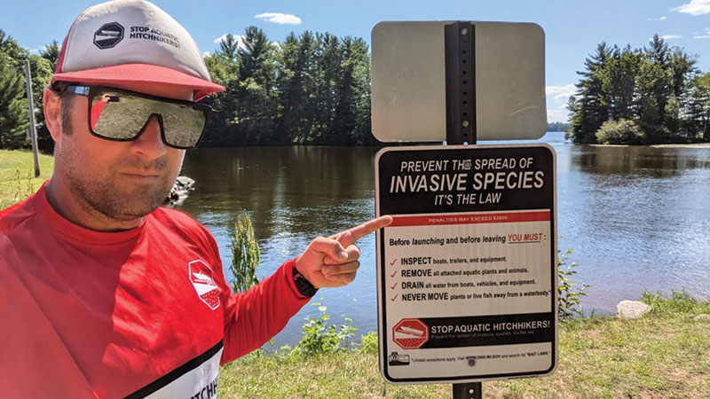 A man in reflective sunglasses wearing a Stop Aquatic Hitchikers shirt points to a sign telling boaters to remove invasive species from their boats.