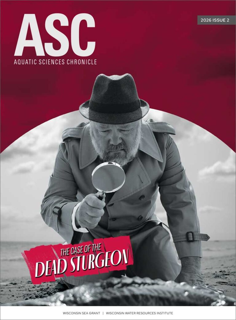 Aquatic Sciences Chronicle 2026 Issue 2 cover featuring a black-and-white image of a man wearing a hat and trenchcoat kneeling on a beach using a magnifying glass to look down at a dead fish.