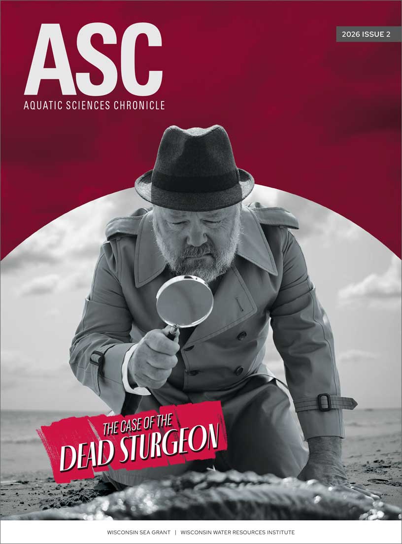 Aquatic Sciences Chronicle 2026 Issue 2 cover featuring a black-and-white image of a man wearing a hat and trenchcoat kneeling on a beach using a magnifying glass to look down at a dead fish.